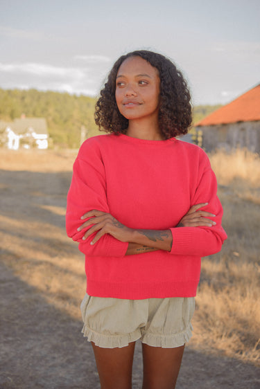 Woman standing outdoors in a field, wearing a red crewneck sweater and green striped bloomer-style shorts.