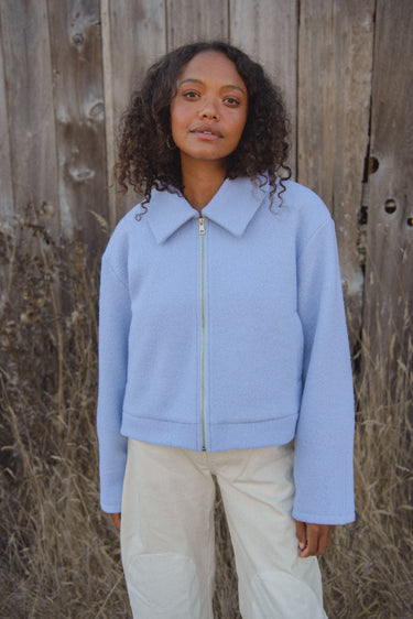 Woman standing in a field outdoors wearing a light blue wool jacket and cream pants.