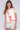 Woman standing in front of a white fabric backdrop wearing a cream vest with red trim over a white short sleeve top, paired with white bloomer-style shorts.