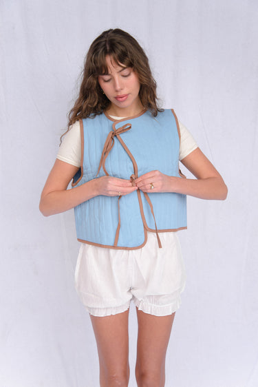 Woman standing in front of a white fabric backdrop wearing a light blue vest with brown trim over a white short sleeve top, paired with white bloomer-style shorts.