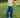 Woman wearing a light yellow sleeveless top and blue casual pants standing in a grassy area with trees in the background.