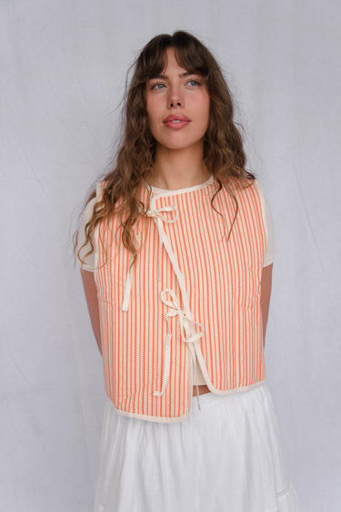 Woman standing in front of a white fabric backdrop wearing a cream and red striped vest with cream trim over a white short sleeve top, paired with a white skirt.