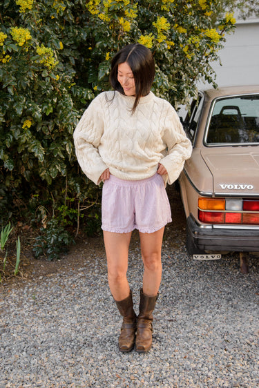 Woman in a cream cable knit sweater and pink plaid boxer-style shorts standing outdoors next to a vintage Volvo car.