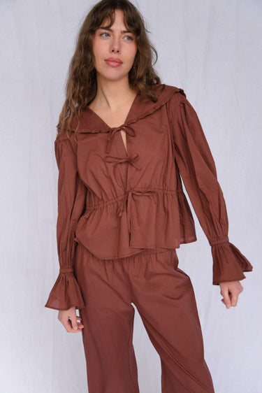 A woman standing in front of a white background wearing a brown top with a sailor-style collar and matching brown pants with an elasticated waistband and cuffed hems. The outfit features drawstrings and the top is tied at the front.