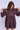 Woman wearing a purple knee-length dress with long sleeves and a ruffled collar on a white fabric backdrop.