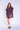 Woman wearing a purple knee-length dress with long sleeves and a ruffled collar on a white fabric backdrop.