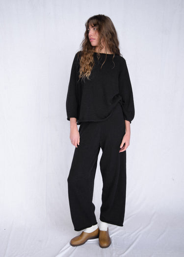 Woman wearing a black, rolled hem sweater and matching black sweater pants on a white fabric background.