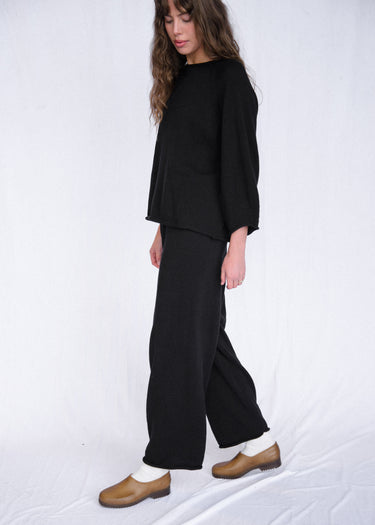 Woman wearing a black, rolled hem sweater and matching black sweater pants on a white fabric background.