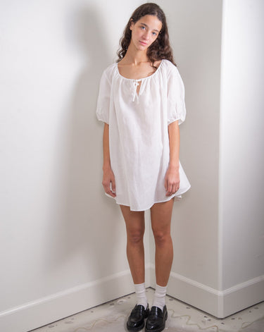 A woman standing against a white wall wearing a white knee-length dress with a tie neckline and short sleeves.