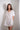 A woman standing against a white wall wearing a white knee-length dress with a tie neckline and short sleeves.