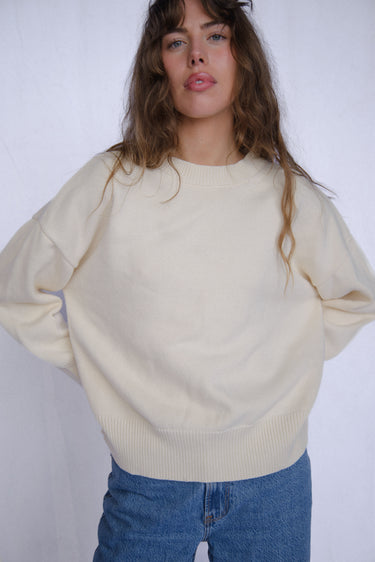 Woman wearing a cream knit sweater and blue jeans against a white background.