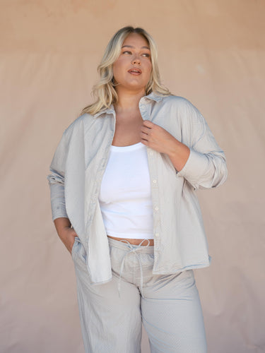 Woman wearing a light gray shirt over a white top and gray pants against a beige background