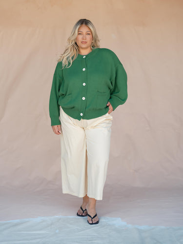 Person wearing a green cardigan and white pants against a beige background