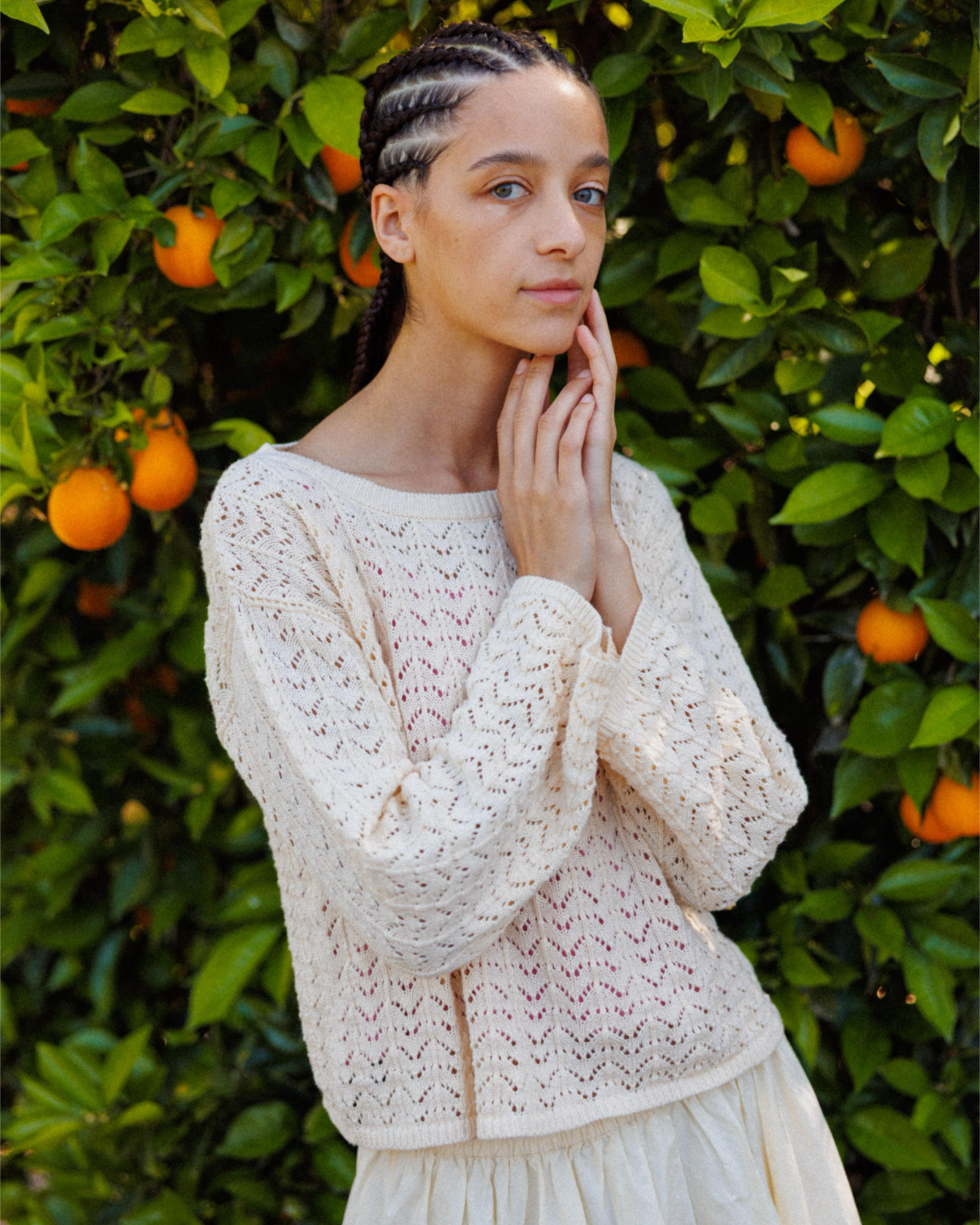 Woman with braids wearing a cream crochet knit sweater in front of an orange grove outdoors.