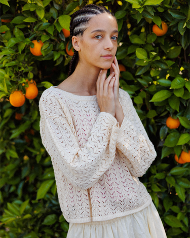 Woman with braids wearing a cream crochet knit sweater in front of an orange grove outdoors.