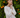 Woman with braids wearing a cream crochet knit sweater in front of an orange grove outdoors.
