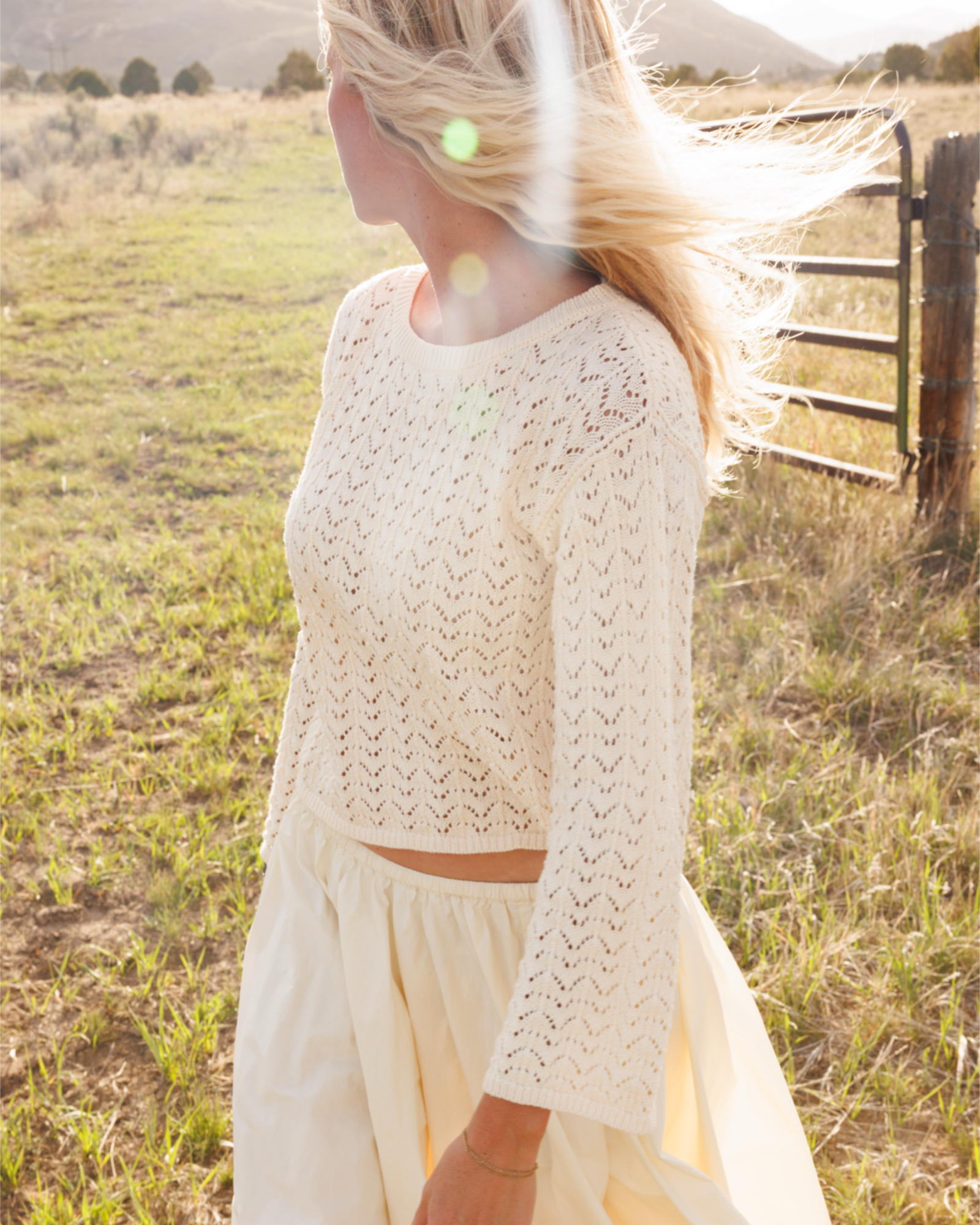 Woman in a crochet knit skirt and cotton skirt standing by a wooden fence in a field.