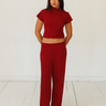 A woman modeling a matching red ribbed two-piece set consisting of a short-sleeve cropped top and wide-leg trousers, standing against a white studio wall and floor.