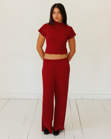 A woman modeling a matching red ribbed two-piece set consisting of a short-sleeve cropped top and wide-leg trousers, standing against a white studio wall and floor.