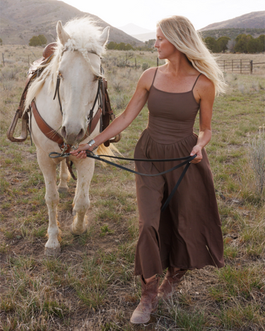 Woman in a brown outfit leading a white horse in a field with mountains in the background.
