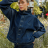 A woman wearing a dark blue denim jacket and matching jeans, posing outdoors in front of a wooden utility pole with green foliage and flowers in the background.