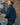 A woman wearing a dark blue denim jacket and matching jeans stands on outdoor concrete stairs next to a brick wall, holding a brown leather bag.