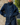 A person wearing an oversized dark blue denim jacket with a high mock neck, front button closure, and patch pockets, standing outdoors against a tree trunk with grass and trees in the background.