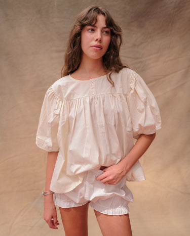 Woman wearing a cream, short-sleeved babydoll-style blouse and white bloomer shorts on a tan fabric background.