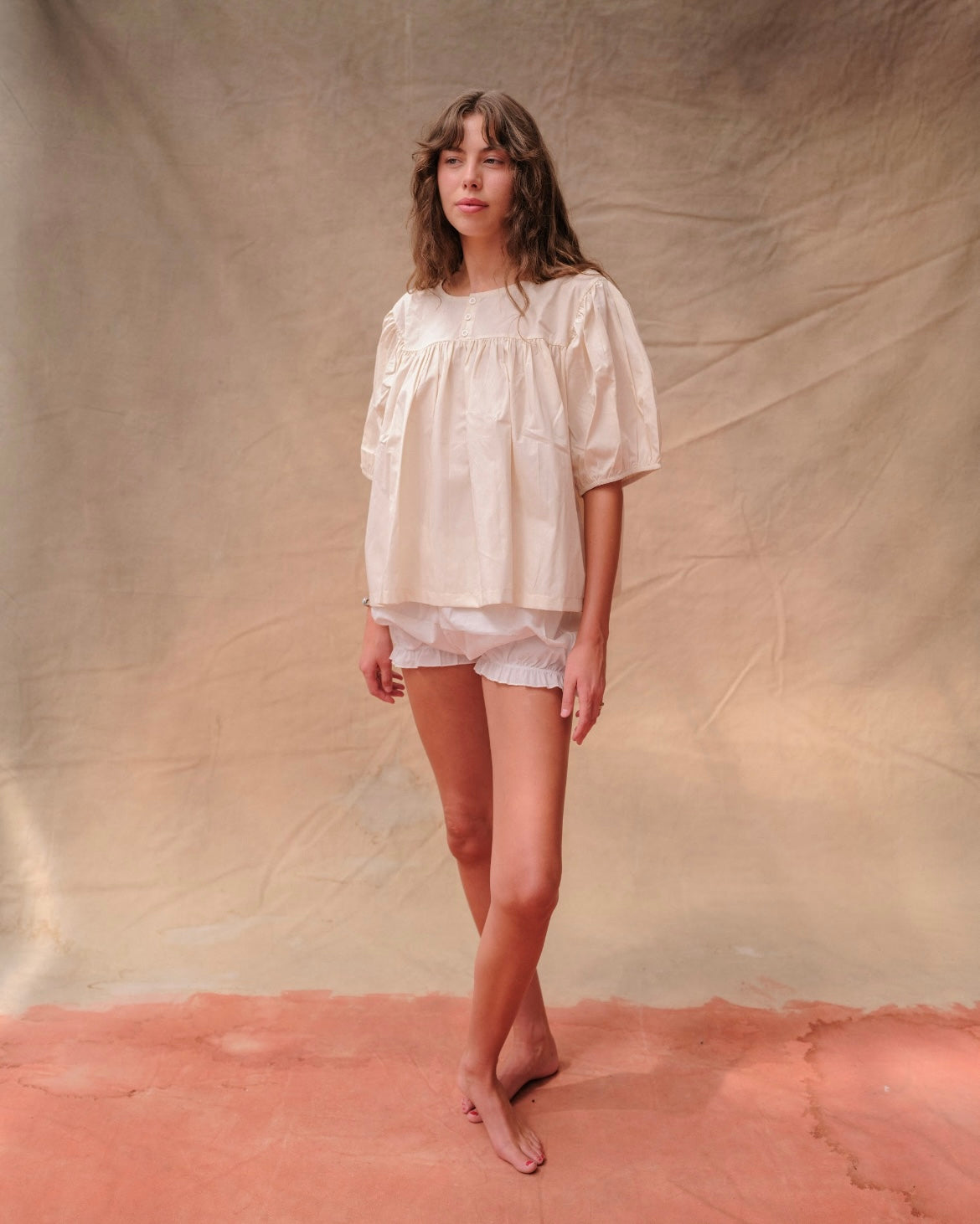 Woman wearing a cream, short-sleeved babydoll-style blouse and white bloomer shorts on a tan fabric background.