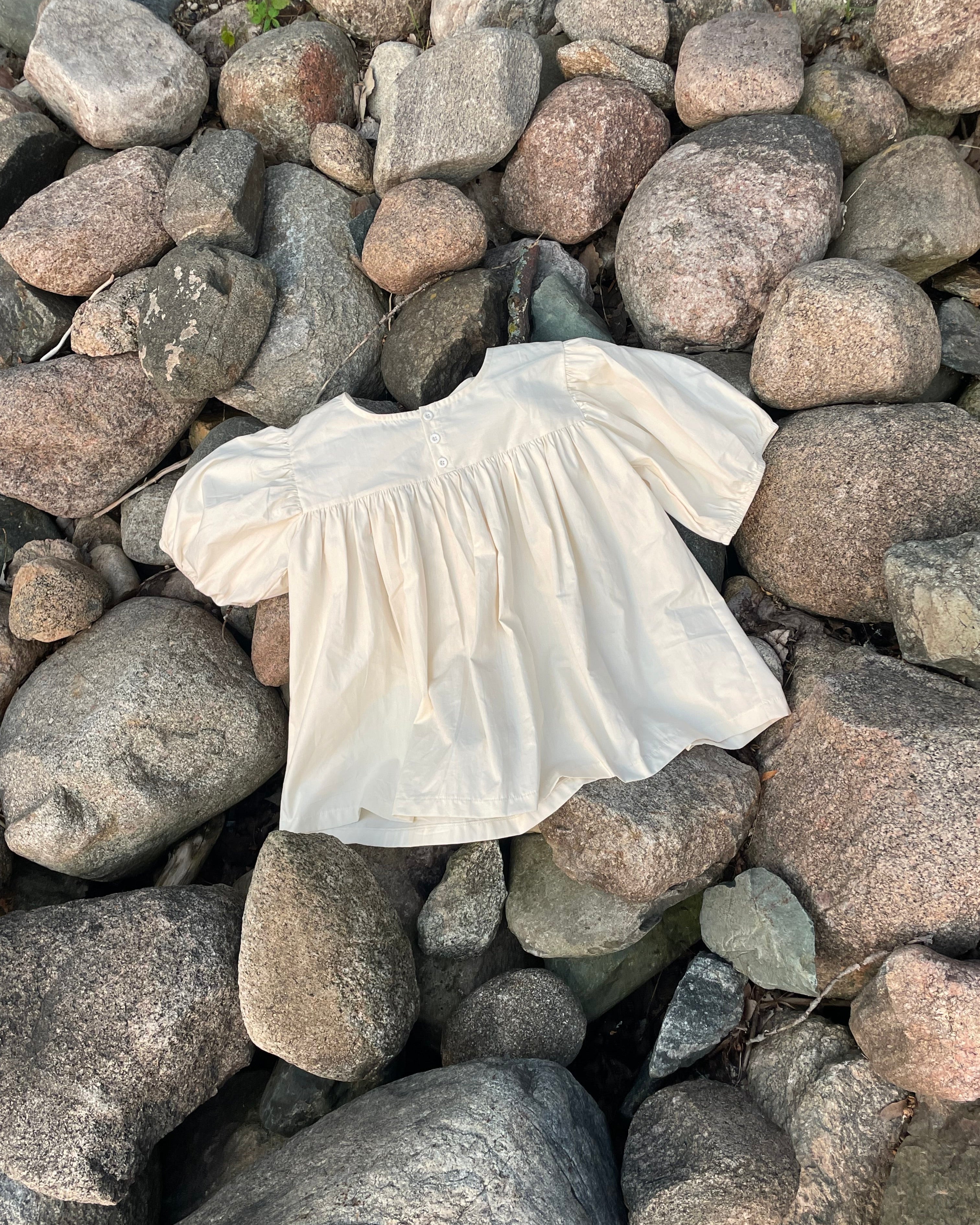Cream babydoll-style blouse with short balloon sleeves laying on top of rocks outside.