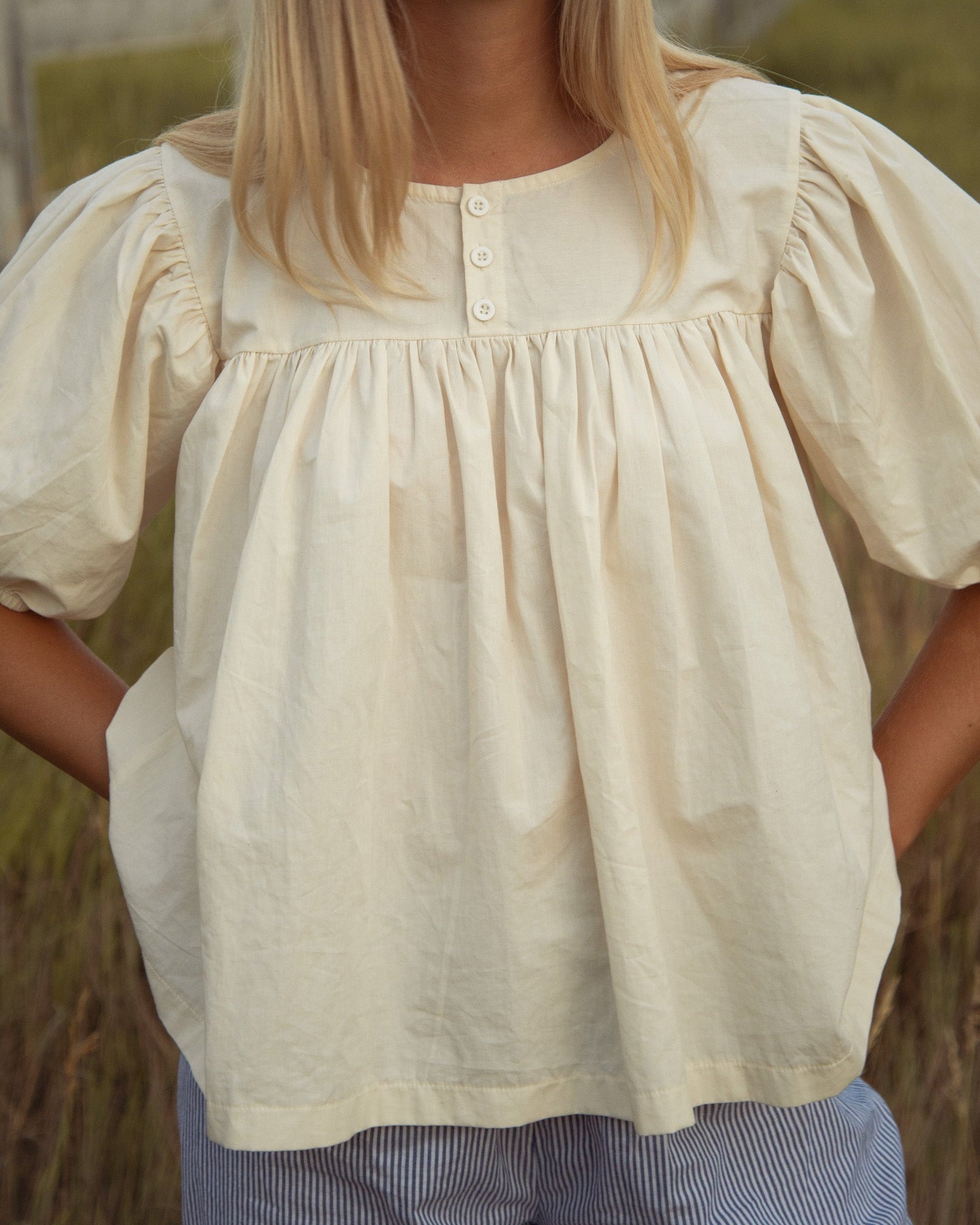 Woman standing in a grassy field outdoors wearing a cream babydoll-style blouse with short balloon sleeves.