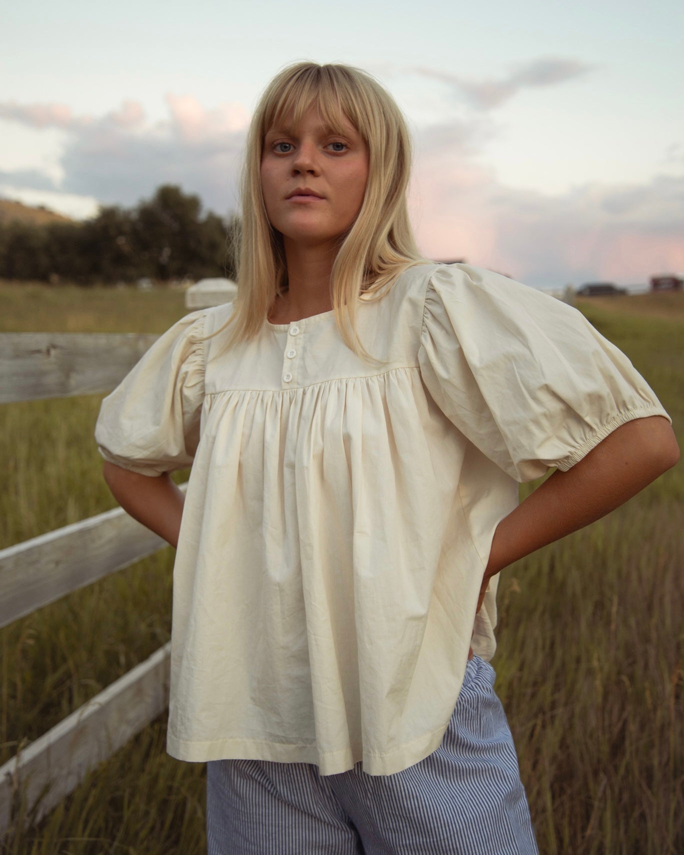 Woman standing in a grassy field outdoors wearing a cream babydoll-style blouse with short balloon sleeves, paired with blue stripe pants.