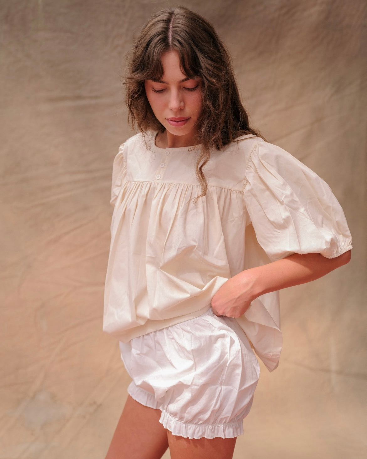 Woman wearing a cream, short-sleeved babydoll-style blouse and white bloomer shorts on a tan fabric background.