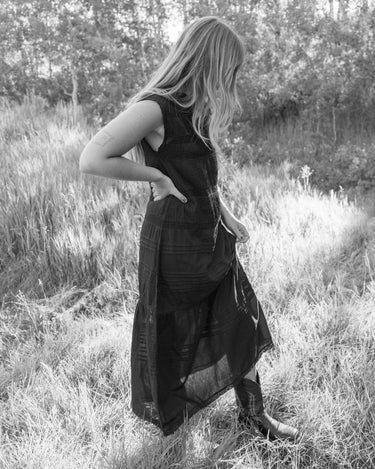 A woman stands in a grassy outdoor field wearing a long dark sleeveless dress and tall boots, photographed in black and white.