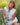 A woman with blonde hair sits in a grassy field, wearing a white sleeveless pleated dress and holding pink wildflowers.