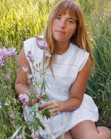 A woman with blonde hair sits in a grassy field, wearing a white sleeveless pleated dress and holding pink wildflowers.