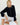 A blonde woman sits barefoot on white wooden floorboards against a white wall, wearing a navy blue long-sleeve button-front cardigan and white ruffled pants.