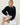 A blonde woman sits barefoot on white wooden floorboards against a white wall, wearing a navy blue long-sleeve button-front cardigan and white ruffled pants.