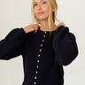 A woman wearing a black ribbed knit cropped sweater with balloon sleeves and a button-down front, paired with white pants, against a plain white background.