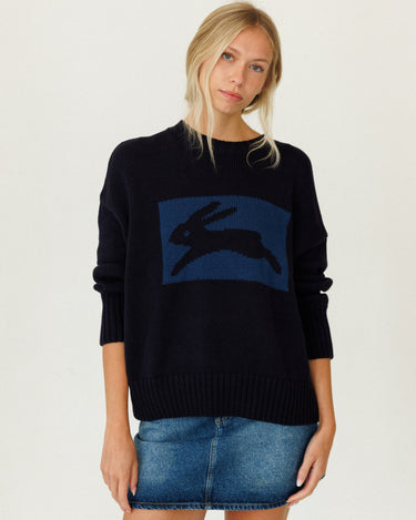 A woman modeling a dark navy blue knit sweater with a large blue rectangular panel on the chest featuring a black rabbit silhouette, paired with a denim mini skirt against a plain light background.