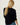 A woman viewed from behind wearing a black knit sweater and blue jeans, with her blonde hair tied back in a ponytail, against a plain off-white background.