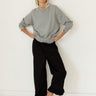A woman posing in a light gray crewneck sweatshirt, black wide-leg cropped pants, and brown lace-up boots against a white wall and floor.