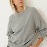 A woman wearing an oversized light gray knit crewneck sweater and black pants, posing against a plain white wall.