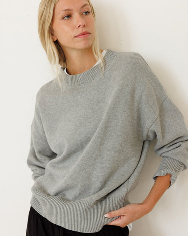 A woman wearing an oversized light gray knit crewneck sweater and black pants, posing against a plain white wall.