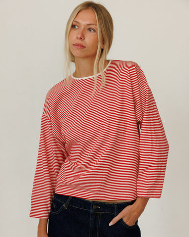 A woman wearing a red and white horizontally striped long-sleeve top with a cream crew neck, paired with dark blue jeans, standing against a plain off-white background.