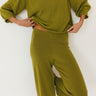 A woman wearing a matching olive green knit lounge set, featuring a long-sleeve sweater and wide-leg pants, posed against a plain white wall.