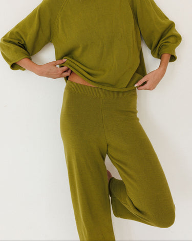 A woman wearing a matching olive green knit lounge set, featuring a long-sleeve sweater and wide-leg pants, posed against a plain white wall.