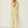 A woman wearing a pale yellow lounge set consisting of a long-sleeve sweater and matching pants, standing barefoot against a white wall on a white wooden floor.