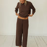 A woman wearing a matching brown lounge set consisting of a long-sleeve cropped top and wide-leg cropped pants, standing barefoot on a white wooden floor against a white wall.