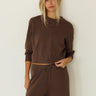 A woman modeling a matching brown lounge set consisting of a cropped long-sleeve sweatshirt and drawstring sweatpants, standing against a plain off-white wall.